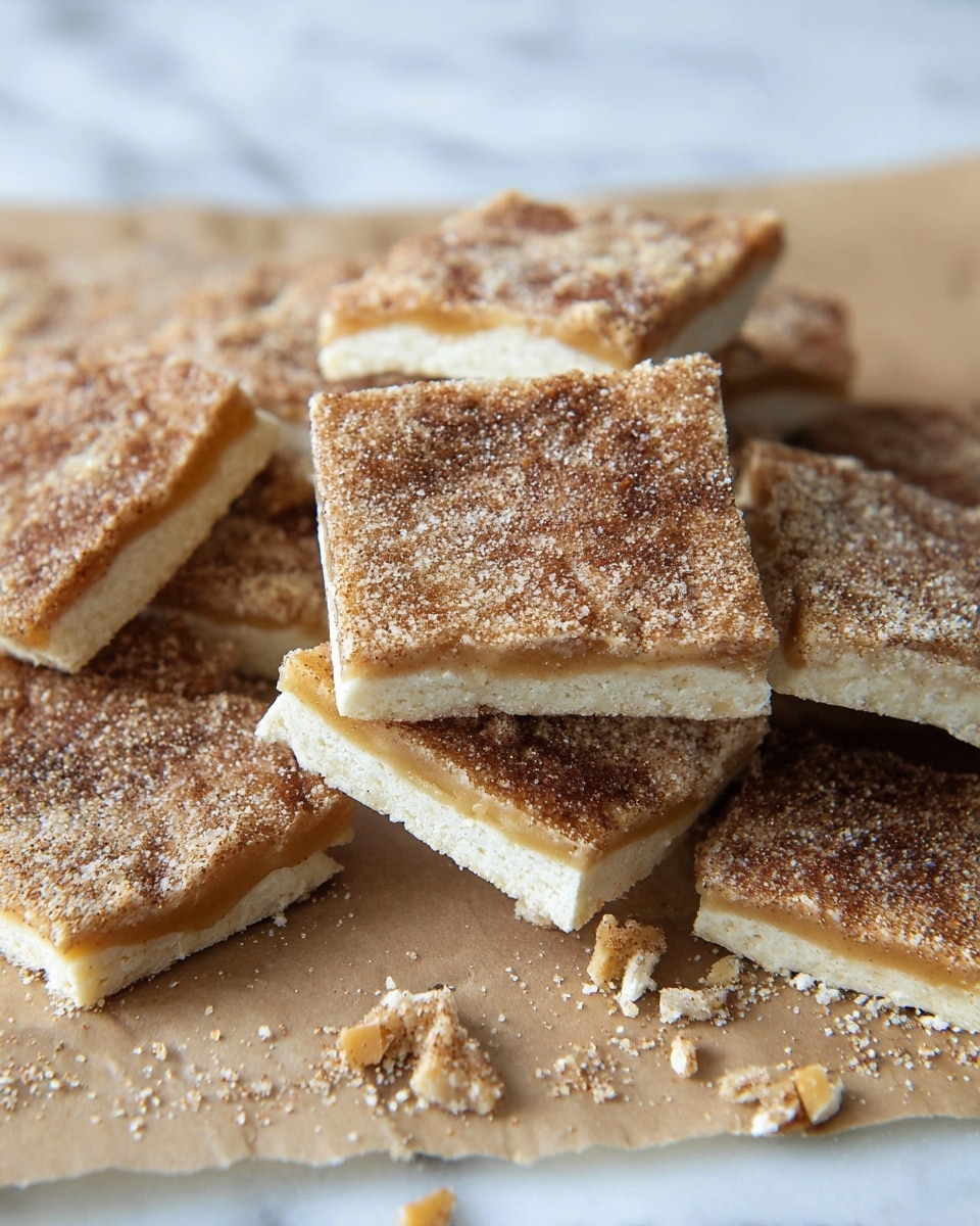 The image shows several square-shaped dessert pieces stacked slightly over each other, with a visible two-layer structure. The top layer is thin and covered with a brown sugar and cinnamon mix that gives a slightly grainy texture and speckled look in light and dark brown shades. The bottom layer is lighter and smooth, resembling a thin dough base with a soft beige color. Some pieces have caramel or toffee filling oozing slightly between the layers, showing a glossy and sticky texture. Small crumbs and broken bits are scattered around on beige parchment paper set on a white marbled surface. photo taken with an iphone --ar 4:5 --v 7