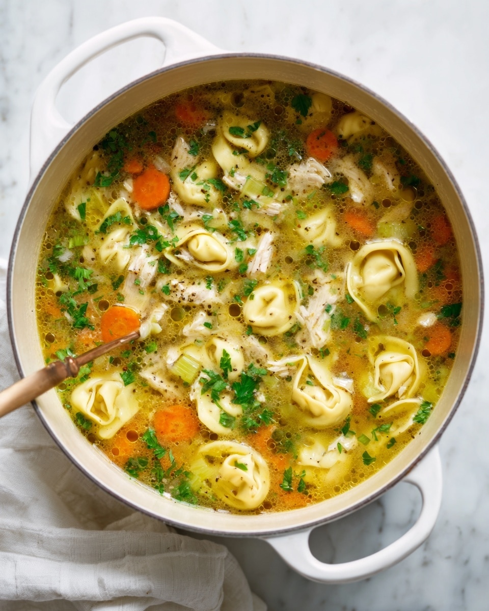 A white pot filled with clear broth soup containing multiple layers of ingredients: the base layer is a golden-yellow broth with visible tiny oil droplets, followed by green parsley leaves and chopped celery scattered throughout, bright orange carrot slices adding vibrant color, small pale yellow tortellini floating on top showing their folded texture, all mixed evenly with some bits of white chicken pieces. The pot rests on a white marbled surface beside a white cloth napkin. Photo taken with an iphone --ar 4:5 --v 7