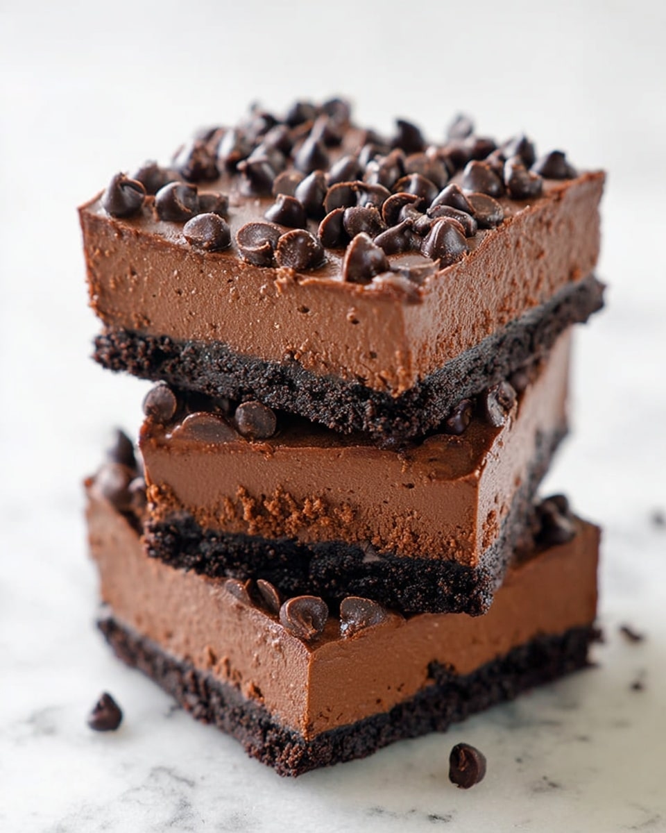 The image shows a stack of three thick chocolate dessert squares on a white marbled surface. Each square has two layers: a dense, dark brown bottom crust and a thick, smooth chocolate mousse layer on top, which is a slightly lighter brown. The mousse is decorated with many small, shiny chocolate chips scattered across the top, adding texture and height. The edges of the squares are cleanly cut, showing the clear separation between the crust and mousse. Pieces of the dessert are also visible around the stack. Photo taken with an iphone --ar 4:5 --v 7