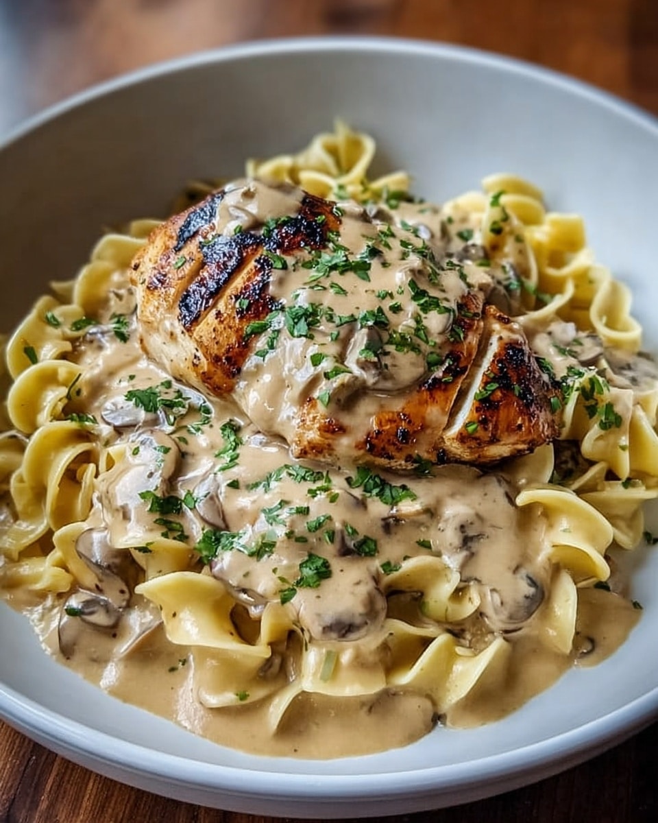 A white bowl is filled with a bed of wide, curly egg noodles in a creamy light beige sauce that pools slightly at the bottom. On top of the noodles lies a grilled chicken breast, golden brown with char marks, covered generously in a thick, smooth beige mushroom cream sauce with visible small mushroom pieces. The dish is garnished with finely chopped fresh green herbs scattered over the sauce and chicken. The bowl is placed on a white marbled surface. Photo taken with an iphone --ar 4:5 --v 7