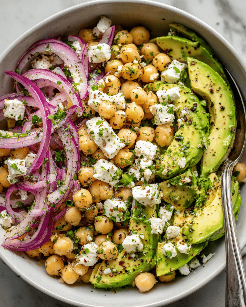 A close-up view of a white bowl filled with a colorful salad consisting of four main layers: the bottom layer is a mix of green avocado slices with a creamy texture, the next layer is beige chickpeas scattered throughout, followed by thin, curved purple-red onion slices adding a sharp contrast, and on top, chunks of white feta cheese and chopped green herbs sprinkled with cracked black pepper and red spices. A silver spoon rests inside the bowl inviting to serve the fresh mix, all placed on a white marbled surface. photo taken with an iphone --ar 4:5 --v 7