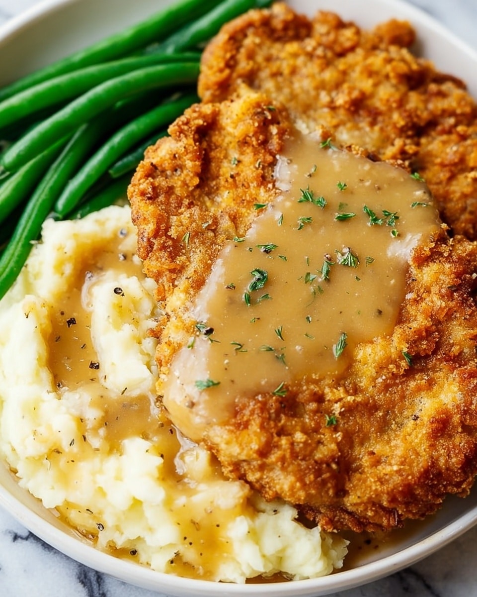 The image shows a white plate with three main layers of food. At the bottom right, there is a rough-textured layer of creamy mashed potatoes, pale yellow in color with some black pepper flakes. On top of the mashed potatoes and spreading toward the center is a smooth layer of light brown gravy with small bits of herbs sprinkled on it. Covering most of the plate is a large, golden-brown crispy fried chicken cutlet with a crunchy, uneven surface full of small craters. To the left side of the plate, fresh green beans add a smooth, shiny texture and a bright green color. The background is a white marbled texture. photo taken with an iphone --ar 4:5 --v 7