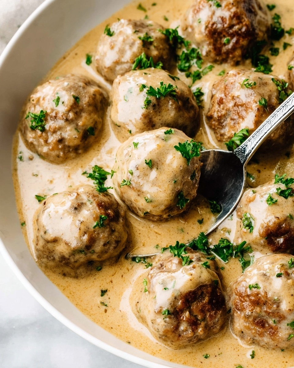 A close-up view of several round meatballs covered in a thick, creamy beige sauce inside a white bowl. The meatballs have a lightly browned and slightly textured surface, partially coated in the smooth sauce that pools around them. Small green parsley leaves are scattered over the meatballs and sauce, adding a fresh pop of color. A silver spoon is partially submerged in the sauce, resting on the right side of the bowl. The bowl is placed on a white marbled surface. photo taken with an iphone --ar 4:5 --v 7