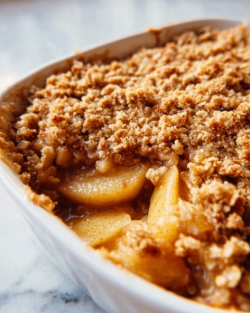 The image shows a close-up of a baked apple dish in a white casserole dish. The bottom layer has soft, sliced apples soaked in a light brown sauce, visible through a gap where a piece is missing. On top, there is a crumbly, golden-brown oat topping that looks crunchy and textured. The dish is placed on a white marbled surface. Photo taken with an iphone --ar 4:5 --v 7