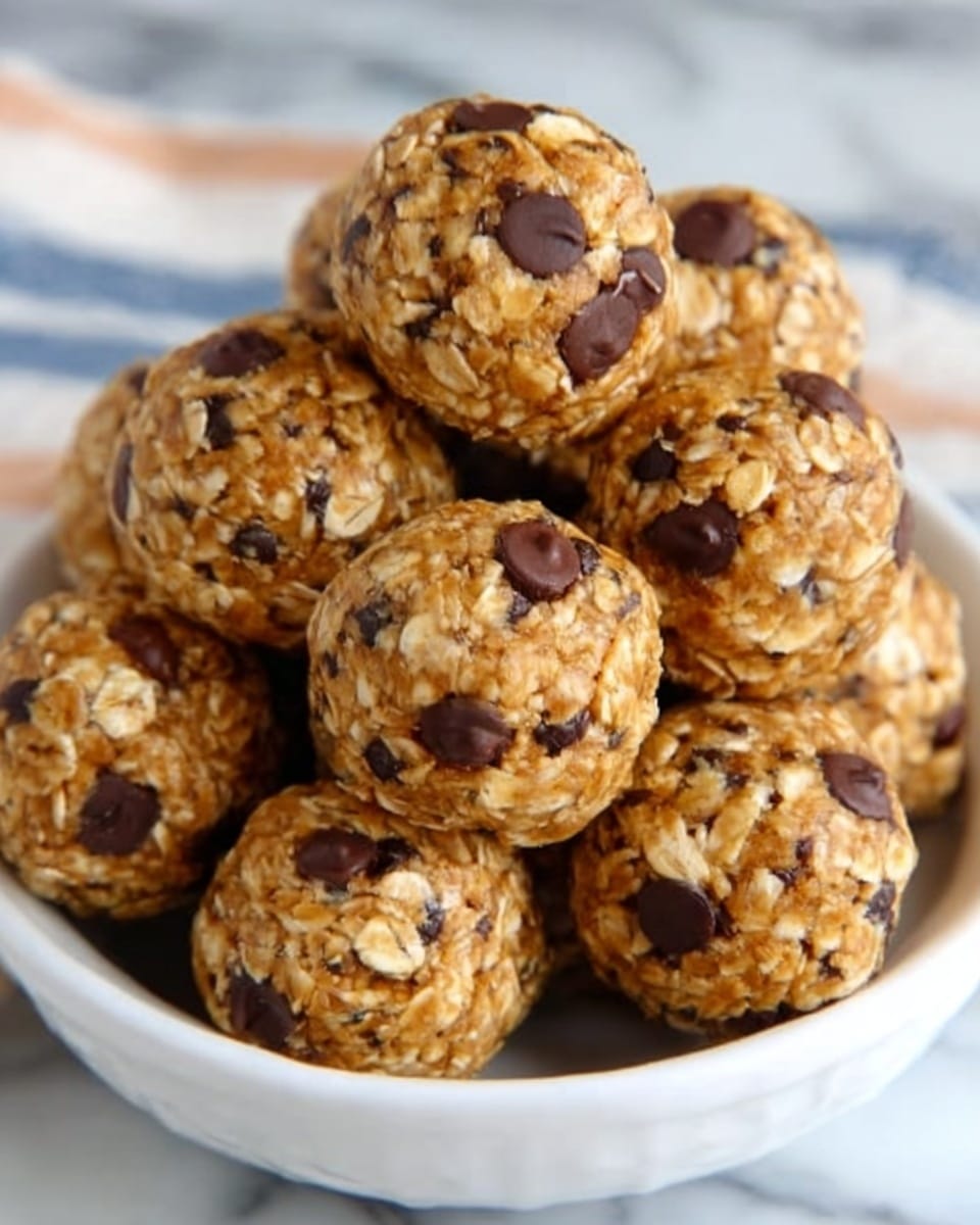 A small white bowl is filled with round energy balls stacked in a pyramid shape. Each ball has a rough texture showing oats mixed with dark chocolate chips scattered all over its light brown surface. The balls appear dense and chewy. The scene is on a white marbled surface. photo taken with an iphone --ar 4:5 --v 7