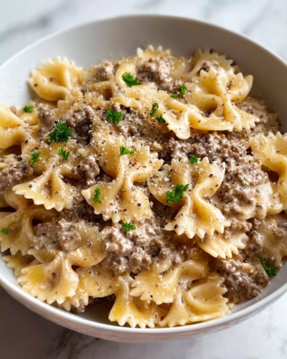 A white bowl filled with farfalle pasta mixed with a creamy sauce and ground beef. The farfalle pasta is light yellow with a slightly glossy texture, layered with browned ground beef scattered evenly throughout. The sauce looks smooth and creamy, coating the pasta and beef, with small flecks of black pepper on top. Garnished with a few small green parsley leaves, adding a fresh color contrast. The background shows a white marbled surface. Photo taken with an iphone --ar 4:5 --v 7