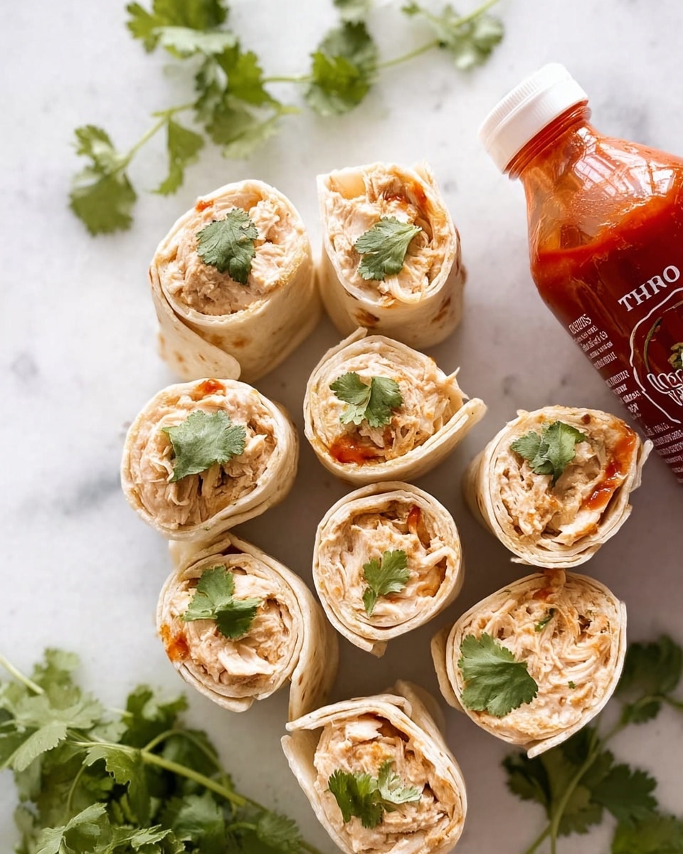 The image shows eight small tortilla pinwheel wraps arranged closely on a white marbled surface. Each wrap has a soft light beige tortilla exterior rolled tightly around a creamy, light tan chicken filling with a slightly rough texture. Small fresh cilantro leaves are scattered both inside the wraps and on top, adding a touch of green color that contrasts with the light beige and tan tones. To the side, there are fresh cilantro sprigs and a white-capped bottle of red hot sauce with visible chili seeds inside, adding a bright red color element. The photo is taken with an iphone --ar 4:5 --v 7