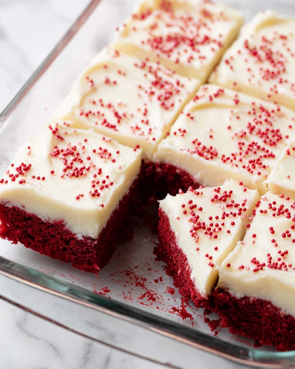 The image shows a glass baking dish with eight cut square pieces of red velvet cake topped with a smooth, thick layer of cream cheese frosting. The frosting has a soft, creamy white color with gentle swirls on the surface, and each cake square is decorated with small round red sprinkles spread evenly. The deep red cake layer underneath the frosting looks moist and dense, giving a strong contrast to the pale frosting. The dish is set on a white marbled surface, and there is some cake crumbs scattered inside the dish around the edges of the cake pieces. Photo taken with an iphone --ar 4:5 --v 7