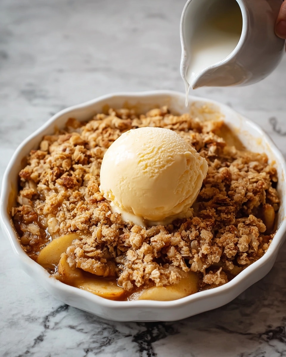 A round white ceramic dish holds a layered apple crumble dessert. The bottom layer is made of cooked apple slices in warm golden-brown syrup, partially visible through the crumbly topping. On top is a thick layer of golden, uneven oat crumble with a rough texture. Sitting in the middle is a single smooth scoop of pale yellow vanilla ice cream, contrasting with the crumbly apple mix below. The dish is placed on a white marbled surface, and a woman's hand holding a white cup is just visible in the background, about to pour something onto the dessert. Photo taken with an iphone --ar 4:5 --v 7