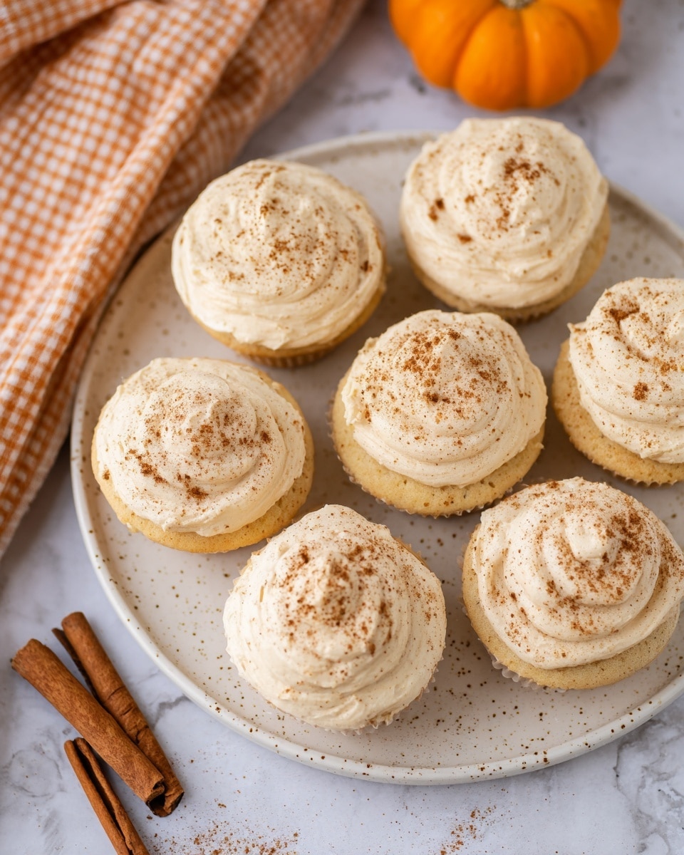 Seven light beige cupcakes with a fluffy and slightly textured frosting sit on a round white speckled plate. Each cupcake is sprinkled with small brown cinnamon powder dots that are unevenly spread on top. The cupcakes have a soft, smooth swirl pattern on their frosting layer, appearing thick and creamy. The plate rests on a white marbled texture surface, with a small bright orange pumpkin in the upper right corner and two cinnamon sticks lying on the lower left, alongside a white and brown checkered cloth peeking in from the top left. photo taken with an iphone --ar 4:5 --v 7