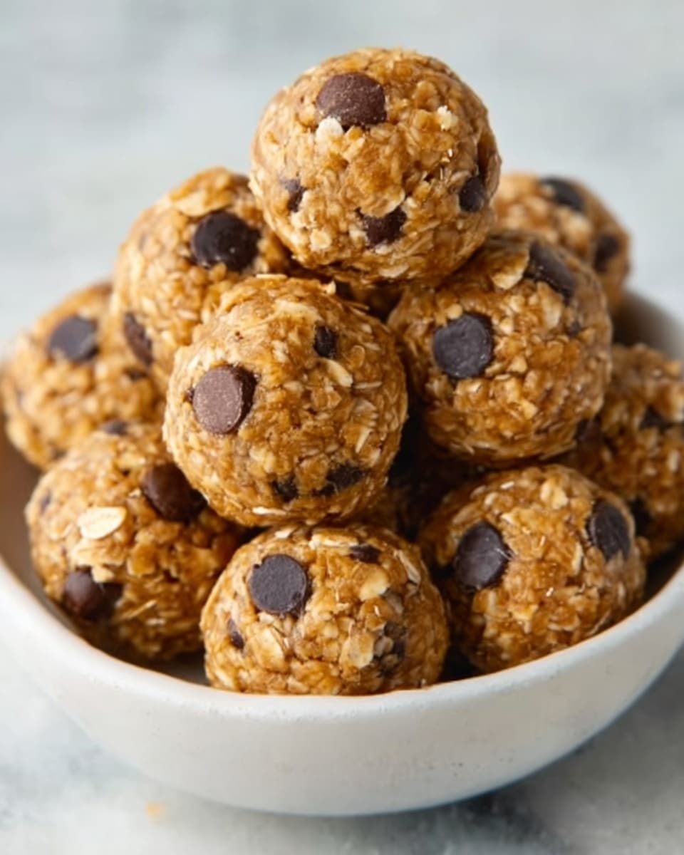 A white bowl filled with a pile of round energy bites, each bite made of a mixture of oats and chocolate chips. The oats give a rough, grainy texture, while the chocolate chips add smooth, dark spots throughout each golden-brown ball. The balls are tightly packed together, creating a small mountain shape. The background shows a white marbled surface. photo taken with an iphone --ar 4:5 --v 7