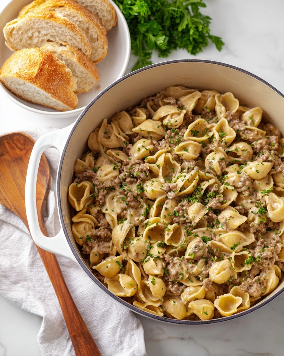 A large white pot filled with creamy pasta mixed with ground meat pieces, topped with small bits of green herbs scattered evenly on the top layer. The pasta shells are pale yellow with a slightly glossy texture, and the meat sauce is light brown and smoothly spread throughout the pasta. To the left of the pot, there is a wooden spoon resting on a white cloth, and behind the pot is a white bowl holding several slices of light brown, crusty bread. Some fresh green parsley leaves are placed around the pot, all set on a white marbled surface. Photo taken with an iphone --ar 4:5 --v 7
