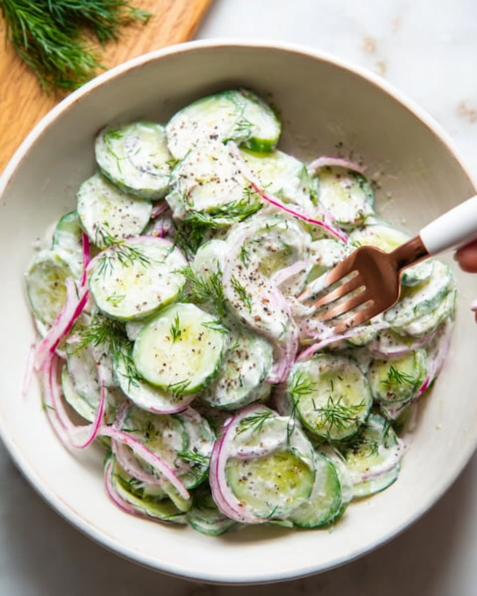 A white bowl filled with cucumber salad layered evenly, showing thinly sliced green cucumbers mixed with thin slices of red onion and fresh dill leaves, all coated in a creamy white dressing with visible black pepper specks. A woman's hand holding a white fork with a copper handle is scooping some salad from the bowl. The bowl is placed on a white marbled surface with some green herbs faintly visible in the background. photo taken with an iphone --ar 4:5 --v 7